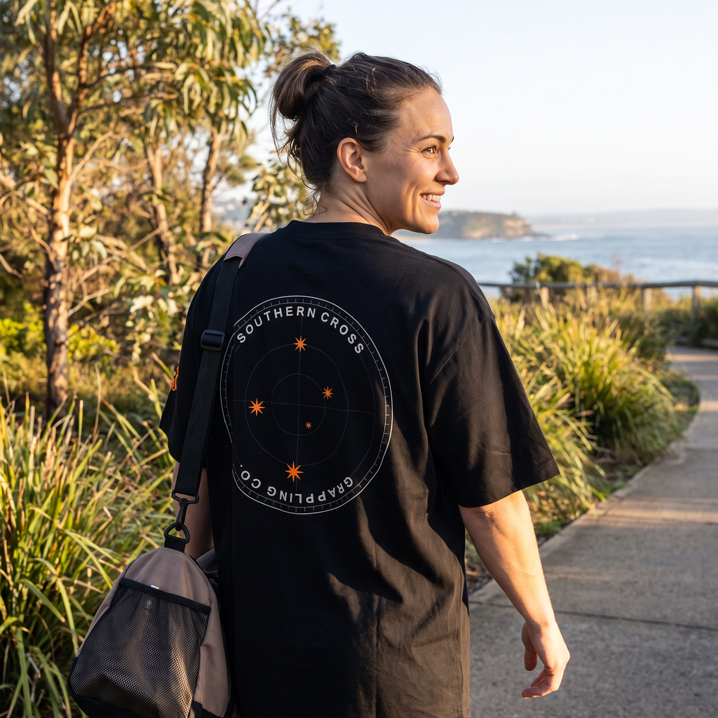 Woman in the Liga Combat Southern Cross Grappling Tee on a coastal park path at sunrise, back emblem visible