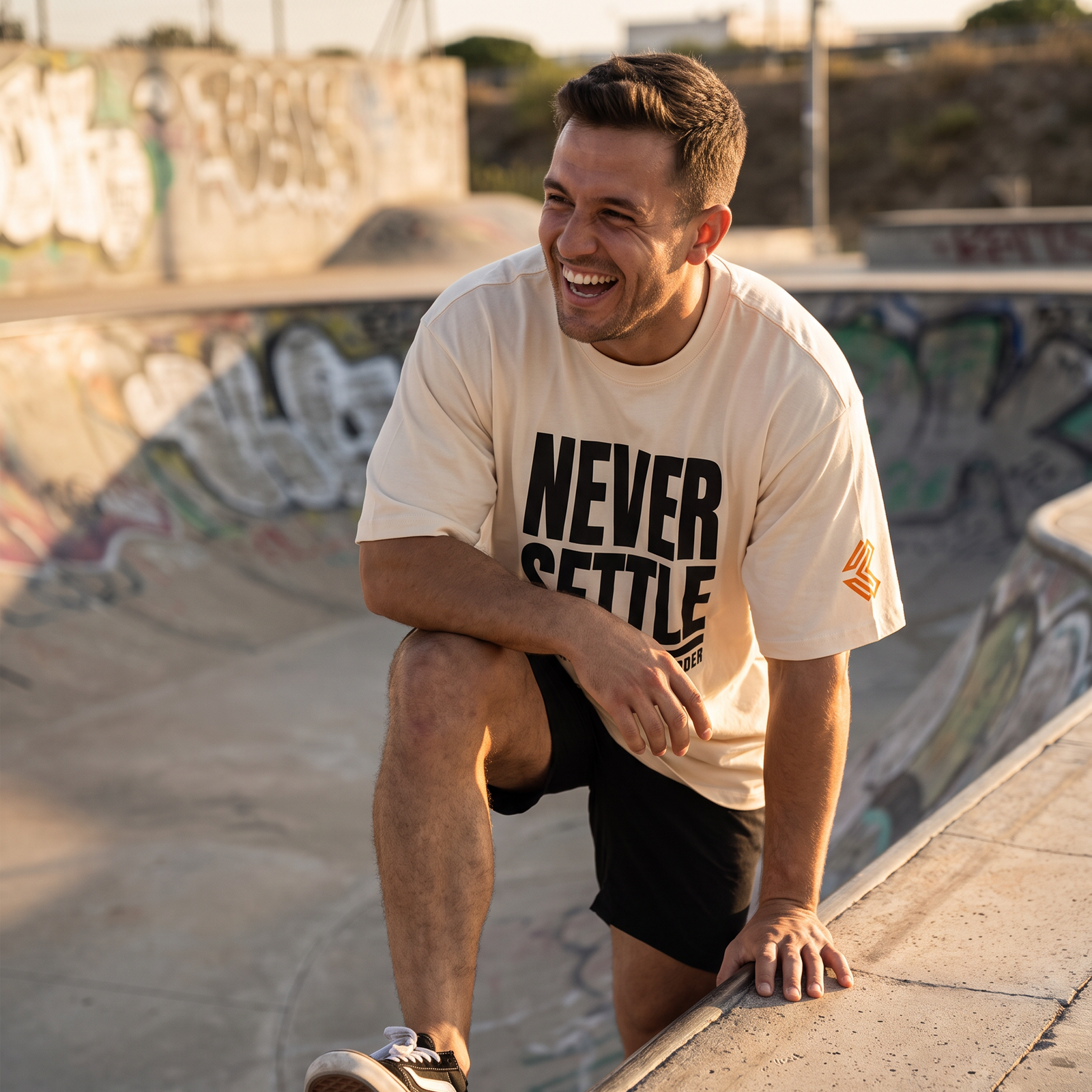 Man in the Liga Combat Never Settle Tee laughing on a skate park ledge at golden hour