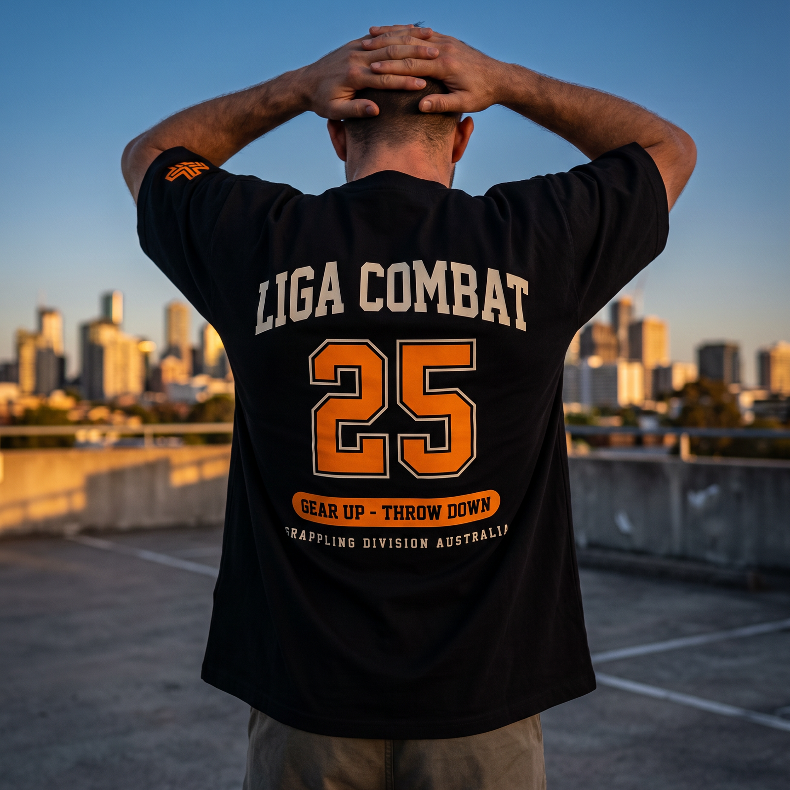 Man in the Liga Combat Varsity 25 Tee on a rooftop at dusk, hands behind his head, city skyline behind